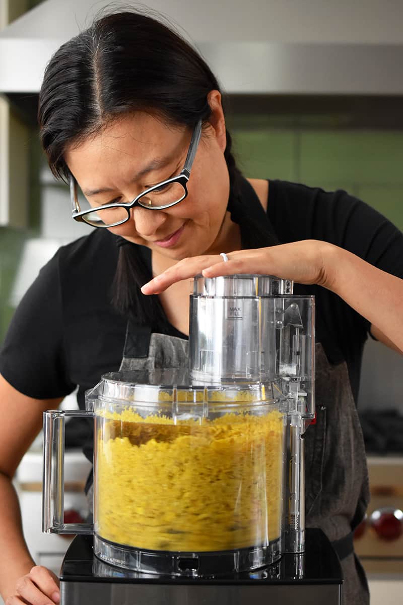 An Asian woman is using a food processor to make crumbs out of plantain chips