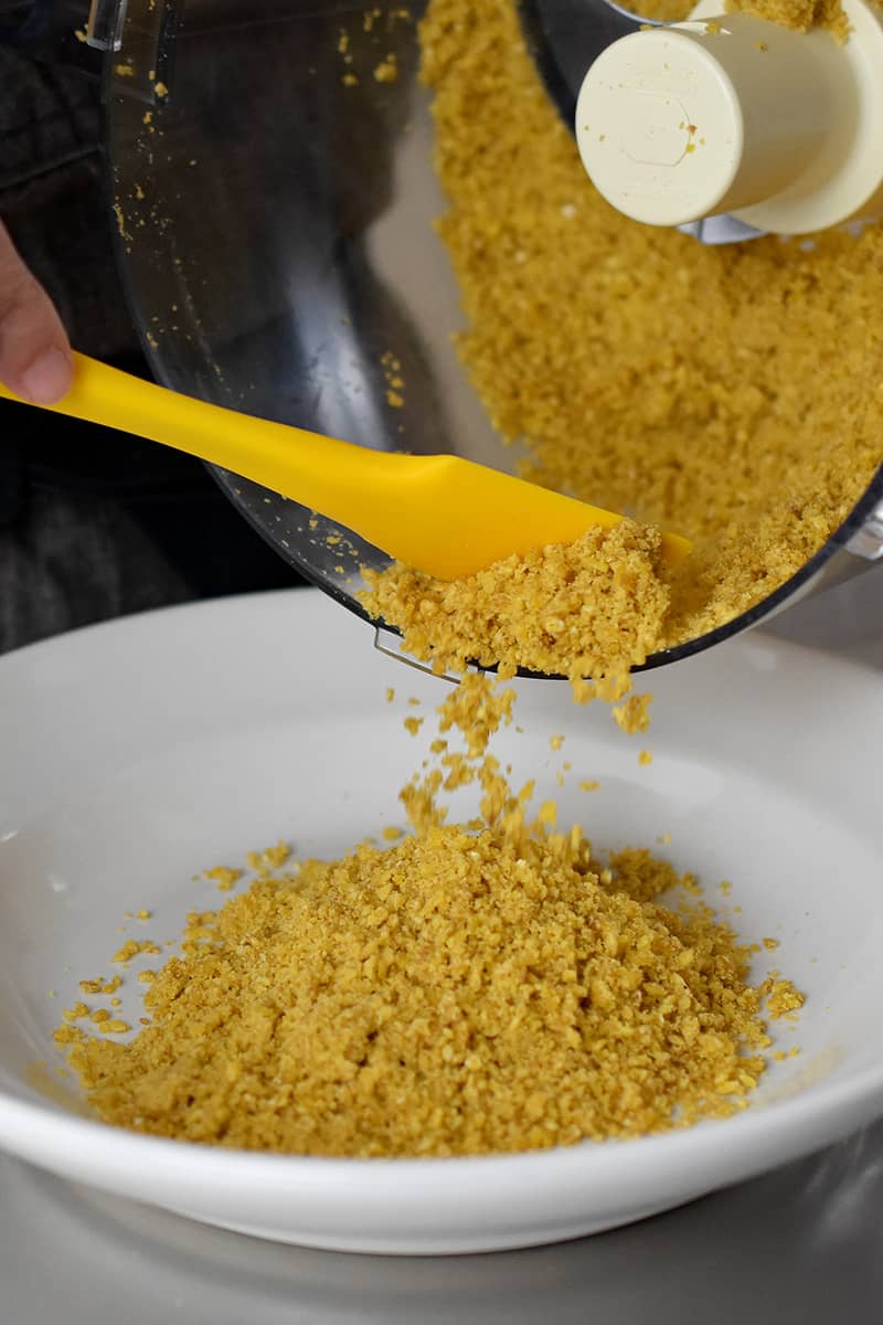 Pouring half of the plantain crumbs from a food processor to a shallow bowl.