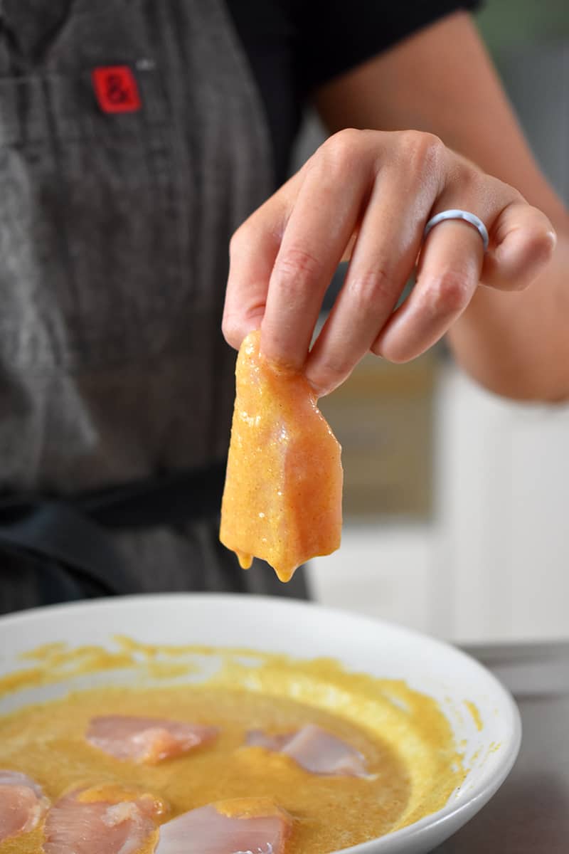 A hand is removing a bite-sized piece of chicken out of a shallow dish filled with an egg-based batter.