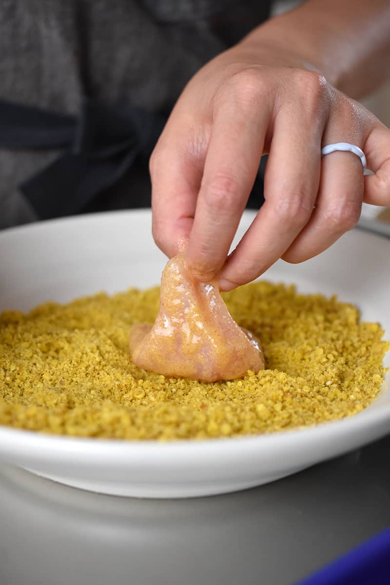 A hand is adding a piece of chicken with a thin layer of batter into a shallow dish filled with plantain crumbs.