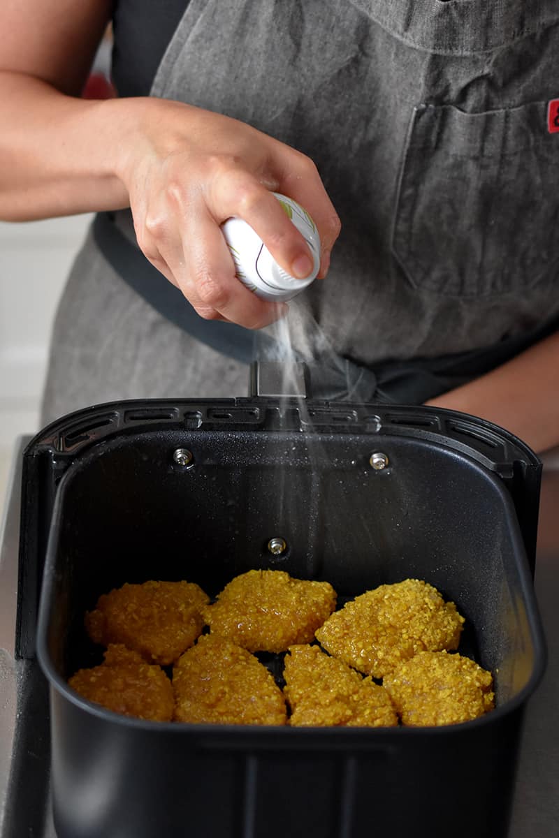 Someone in a gray apron is spraying avocado oil on some chicken nuggets in an air fryer basket.