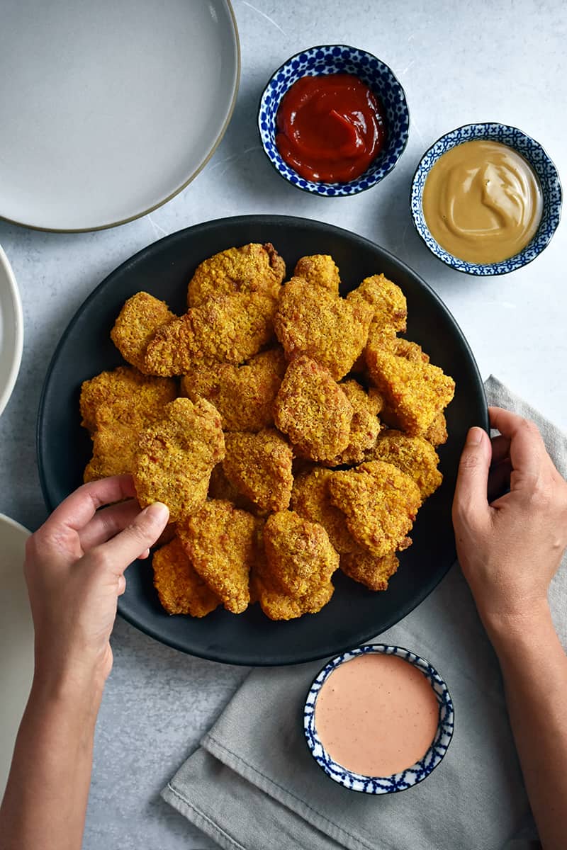 An overhead shot of someone grabbing a chicken nugget from a plate. There are three bowls filled with dipping sauces next to it.
