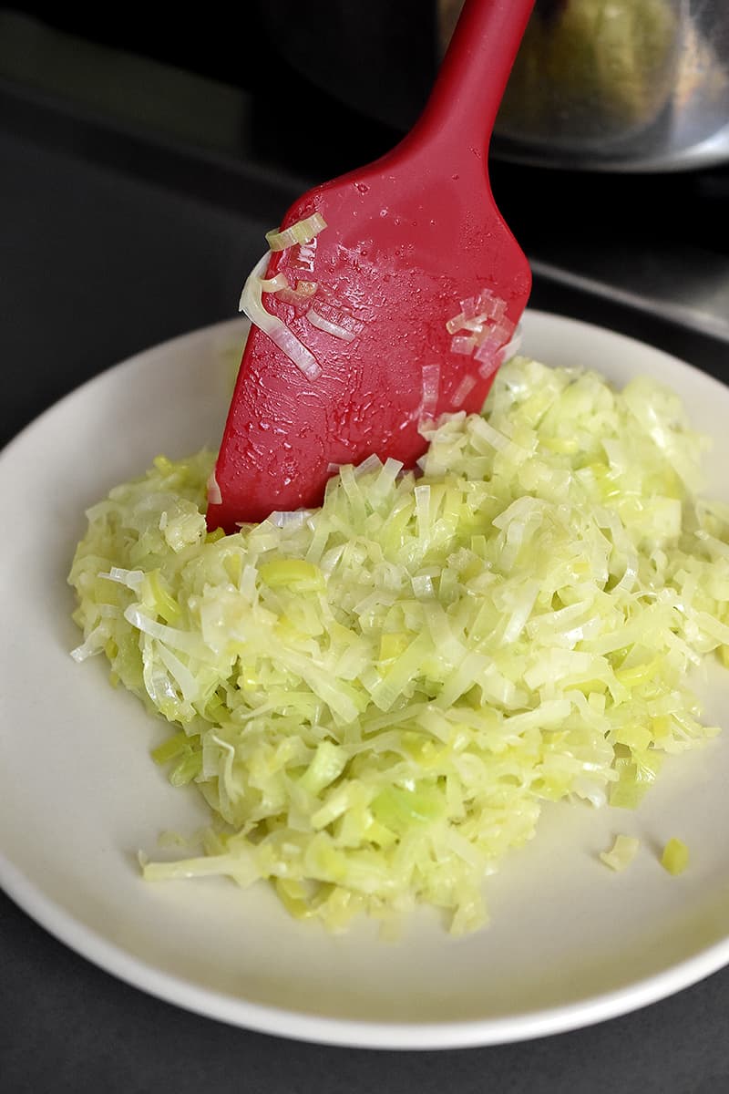 A red spatula is transferring cooked leeks to a plate.