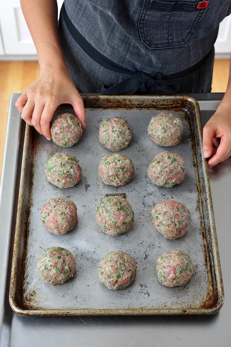 A hand is placing a meatball on a rimmed baking sheet filled with 12 meatballs.