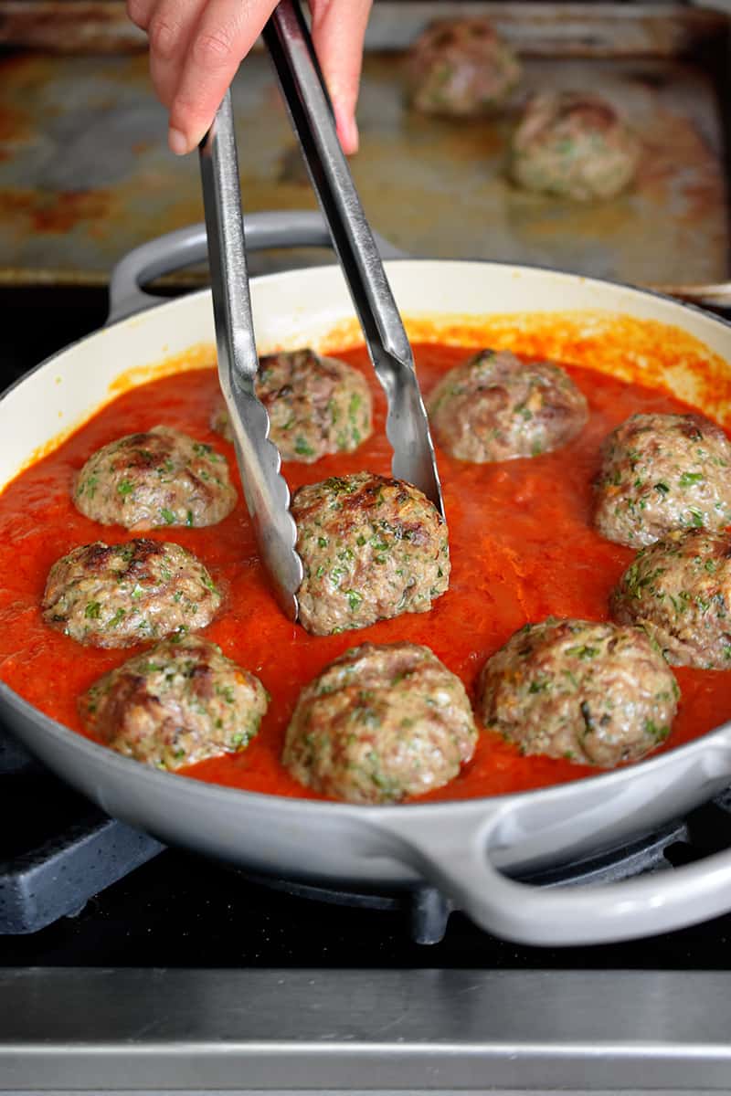 A pair of tongs is adding broiled meatballs to a skillet filled with marinara sauce.