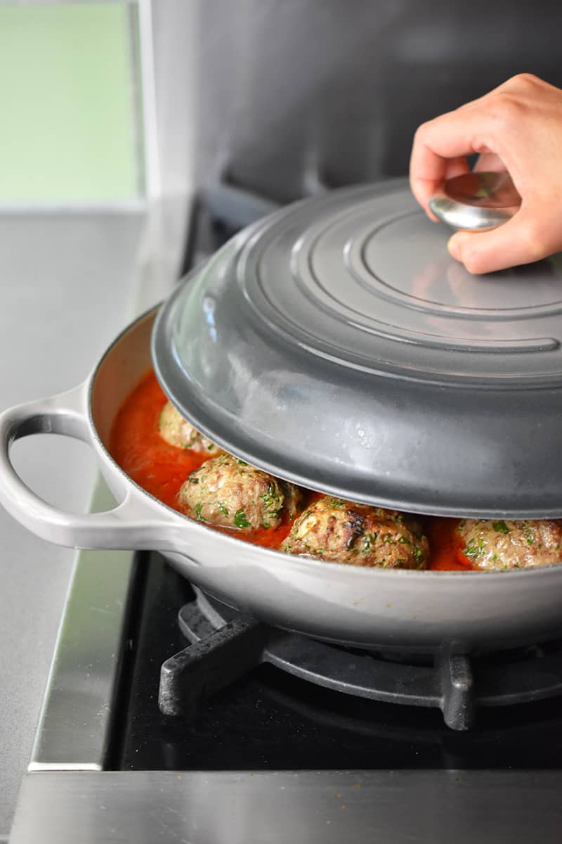 A hand is partially covering a gray Le Creuset skillet filled with Italian meatballs and marinara sauce