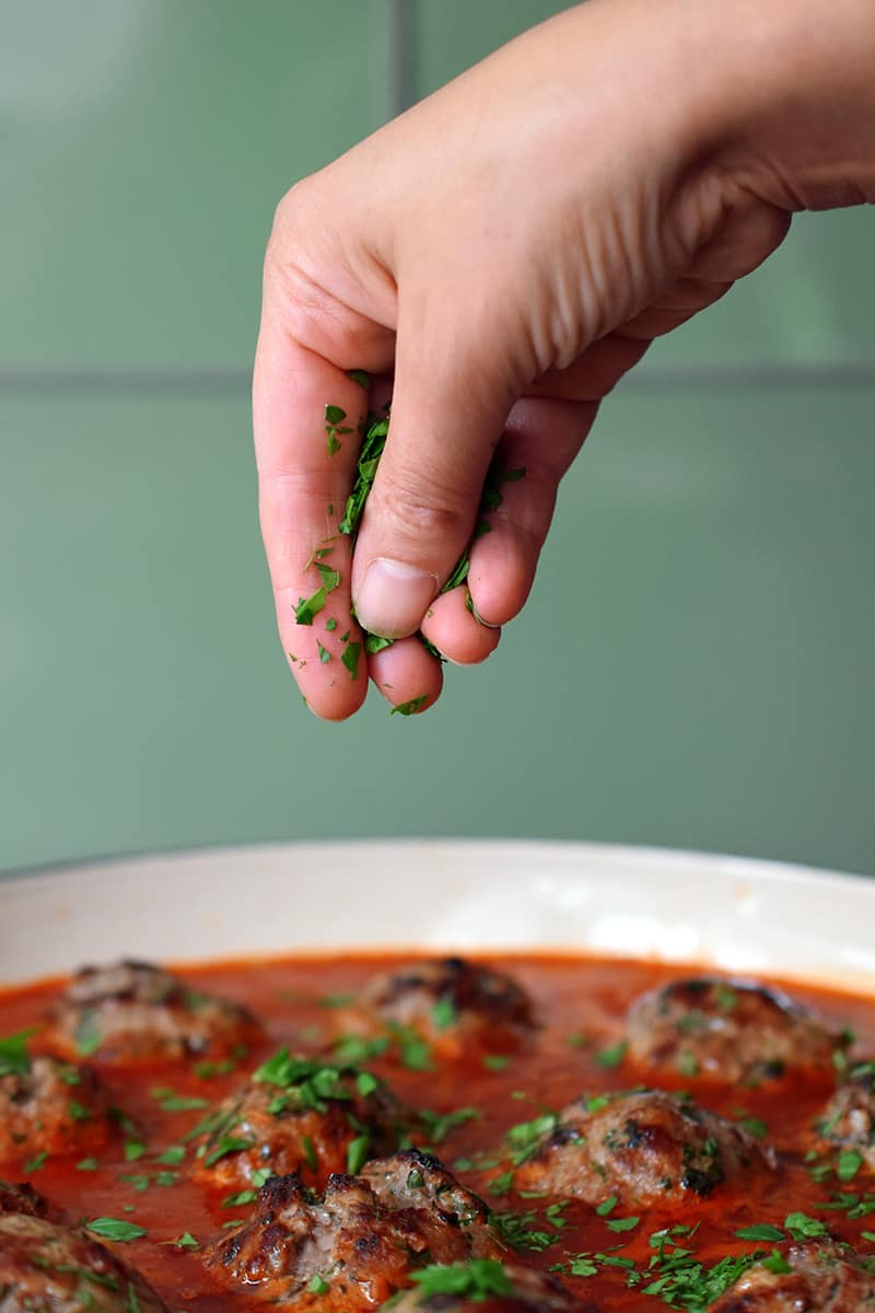 A hand is sprinkling minced Italian parsley onto a skillet filled with Italian meatballs and marinara sauce.