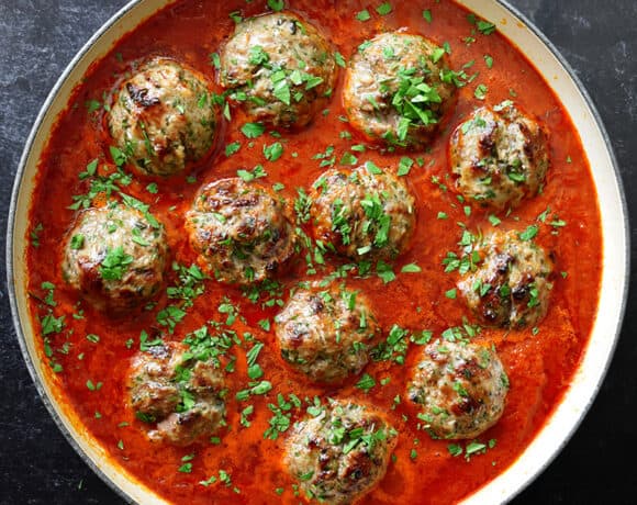 Two hands are holding the handles of an enameled cast iron skillet filled with Italian meatballs in marinara sauce and topped with minced Italian parsley