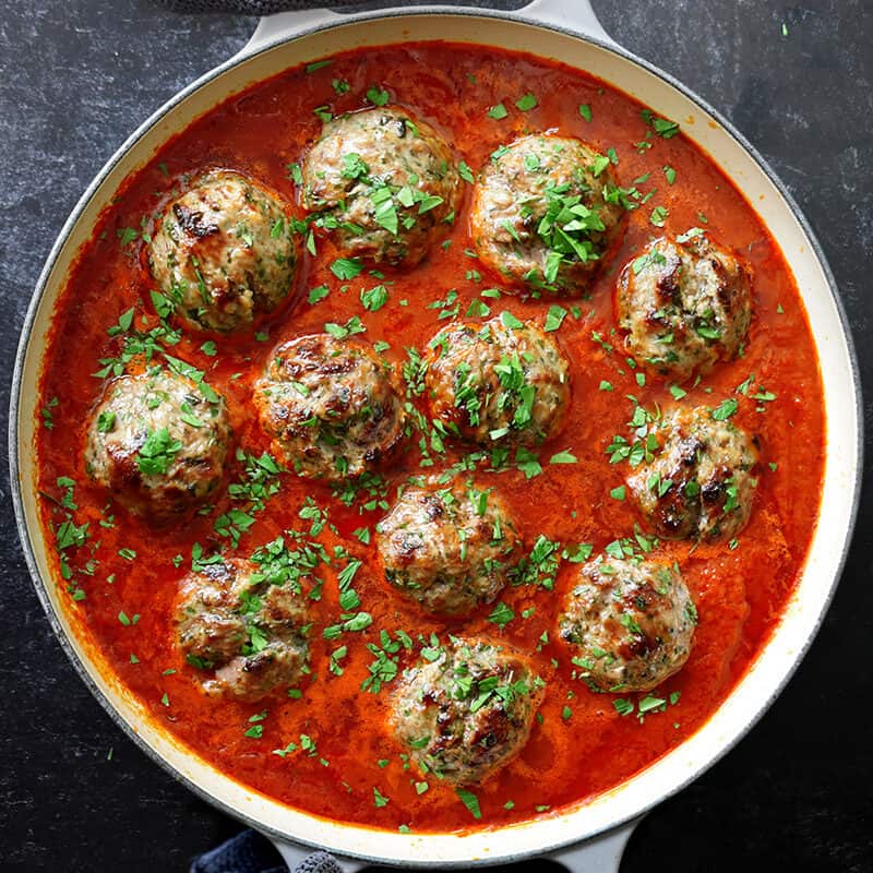 Two hands are holding the handles of an enameled cast iron skillet filled with Italian meatballs in marinara sauce and topped with minced Italian parsley