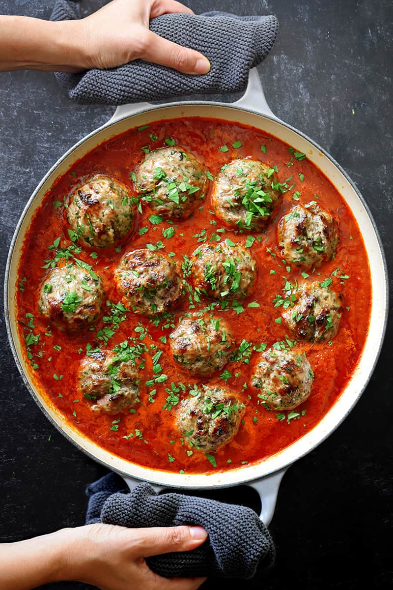 Two hands are holding the handles of an enameled cast iron skillet filled with Italian meatballs in marinara sauce and topped with minced Italian parsley