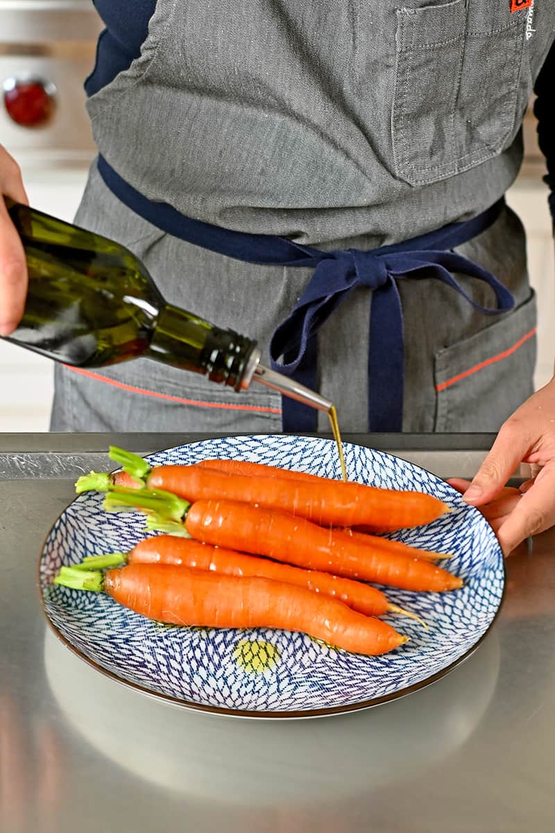 Drizzling olive oil from a bottle onto some carrots on a blue plate.
