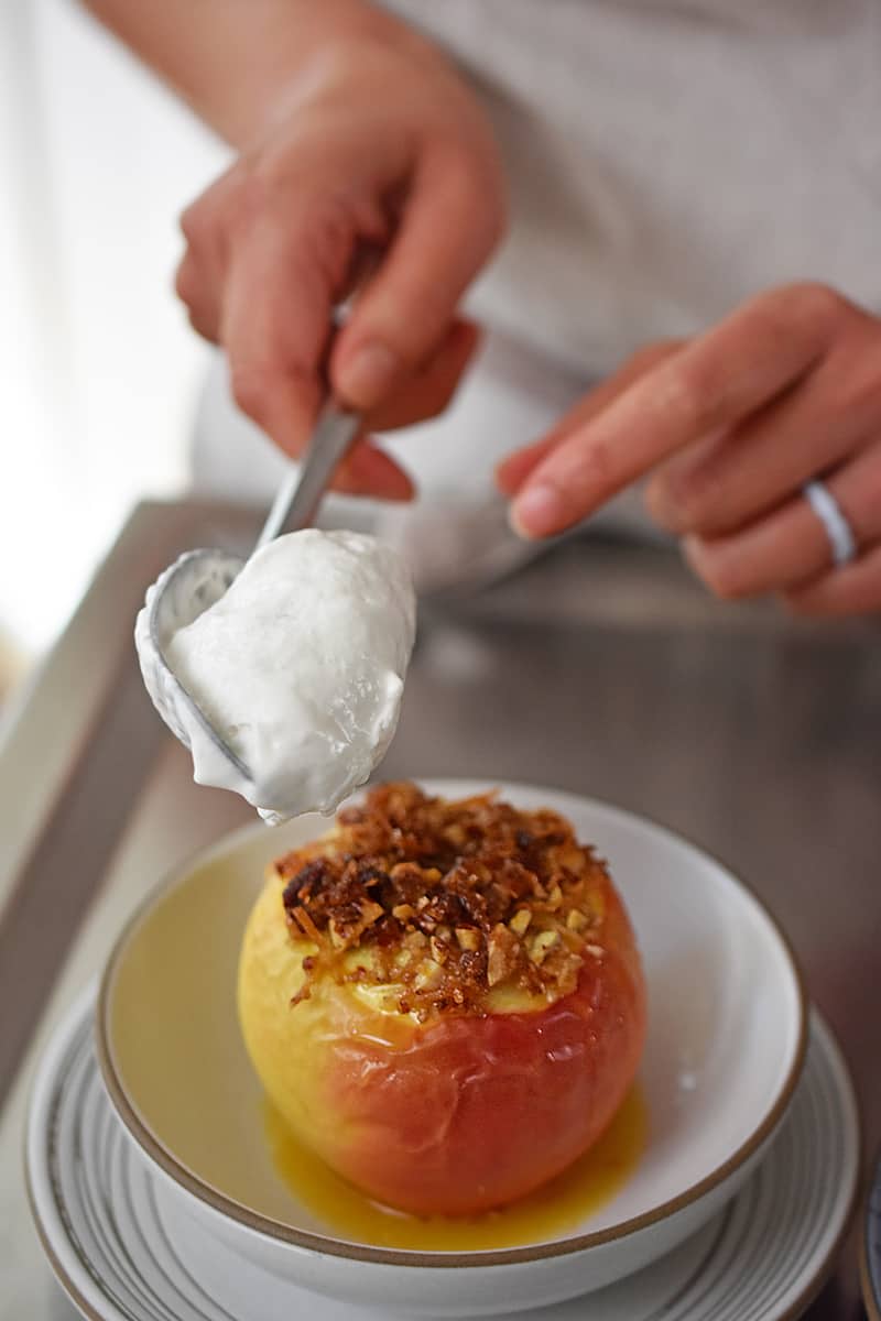 A spoon is adding a dollop of whipped cream to a baked apple in a shallow white bowl.