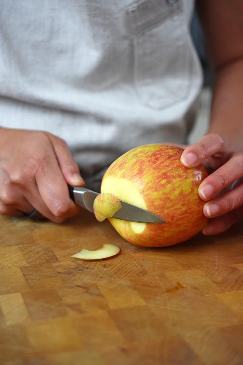 Someone using a paring knifer to trim the bottom of the apple on a wooden cutting board.