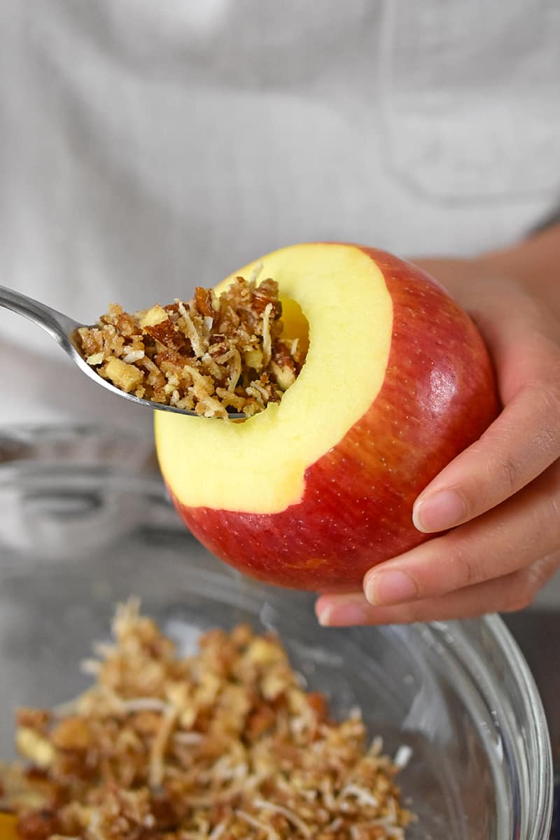 A spoon is filling a cored apple with a paleo and vegan sweet and salty filling.
