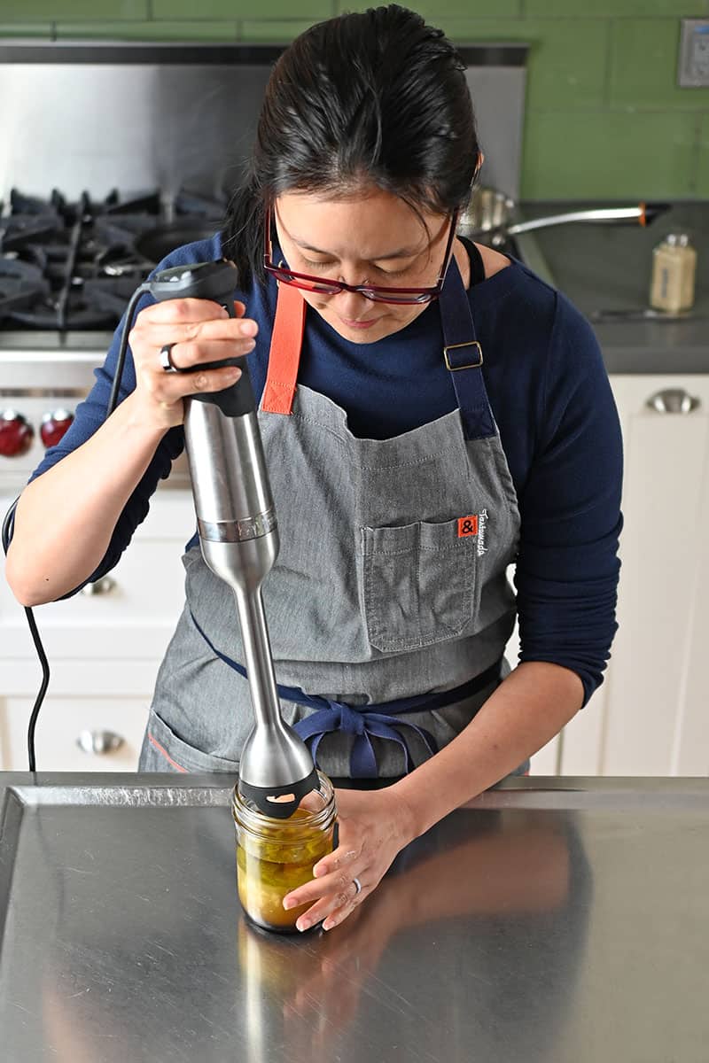 An Asian woman in a gray apron is using an immersion blender to mix sesame ginger dressing in a small mason jar.