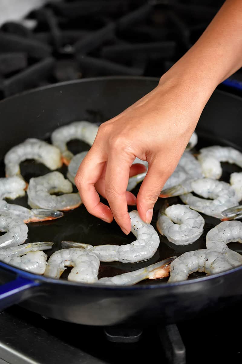 A hand is adding seasoned raw shrimp to a large cast iron skillet on the stovetop.