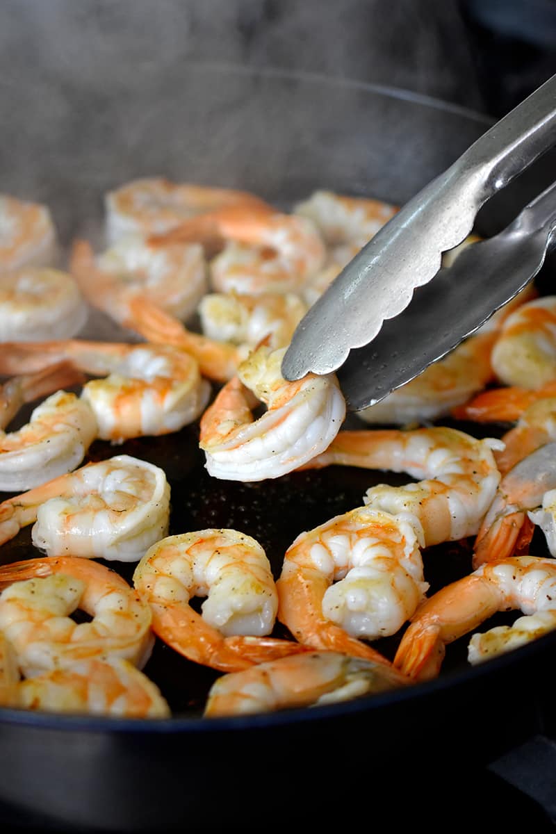 A pair of tongs is flipping shrimp in a large cast iron skillet.