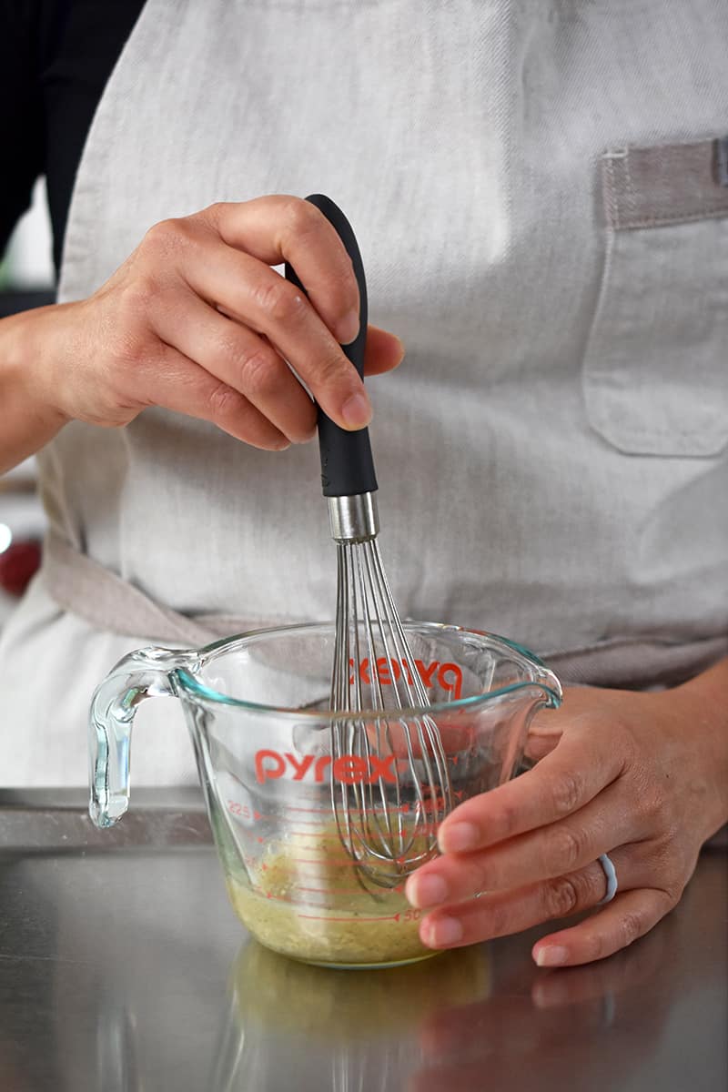 A person in a beige apron is whisking champagne vinaigrette in a liquid measuring cup.
