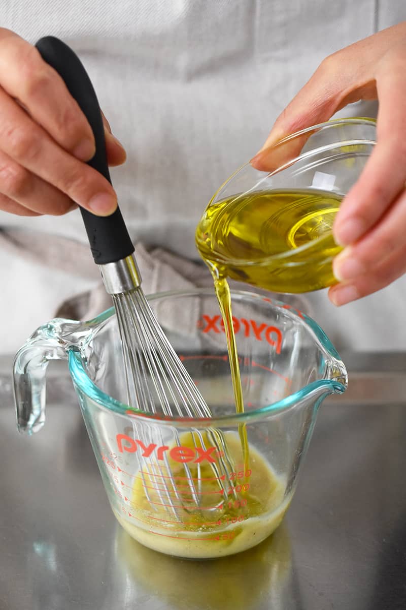 A small bowl of extra virgin olive oil is being drizzled into a measuring cup that is filled with champagne vinaigrette ingredients being whisked.