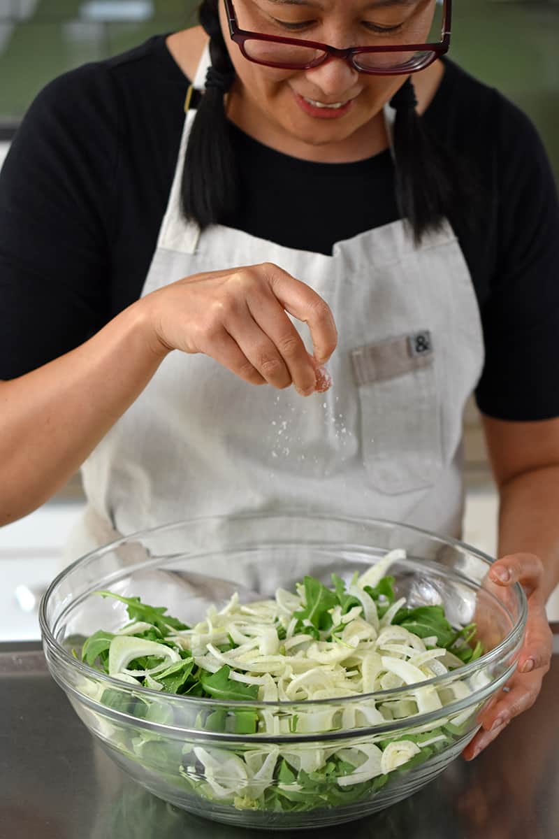An Asian woman in glasses is seasoning some baby arugula and shaved fennel in a large bowl.