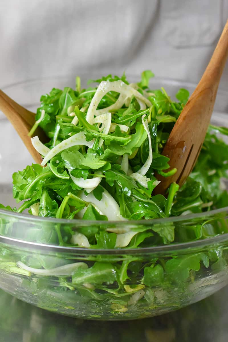 Two wooden spoons are tossing fennel and arugula in a large glass bowl.