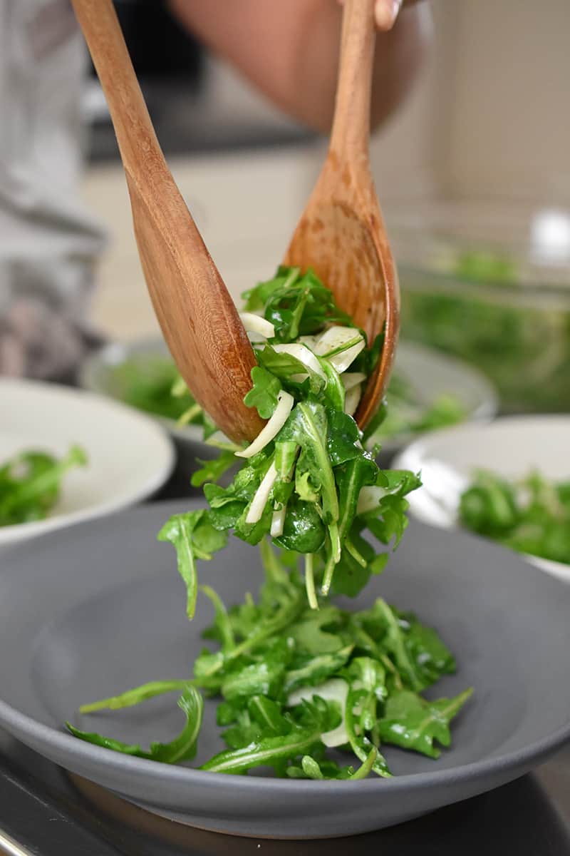 Two wooden spoons are dividing salad greens onto a gray shallow bowl.