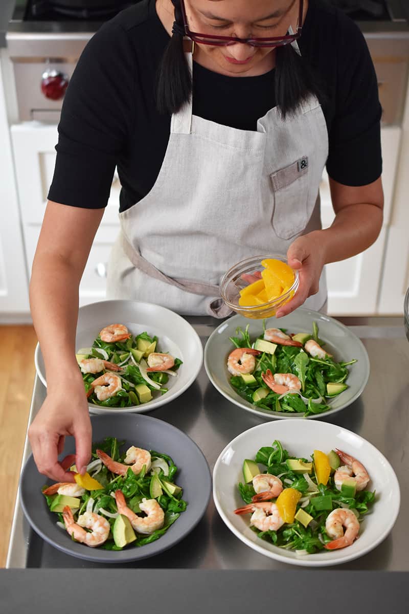 An Asian woman in a beige apron is adding orange segments to four bowls of shrimp and avocado salad