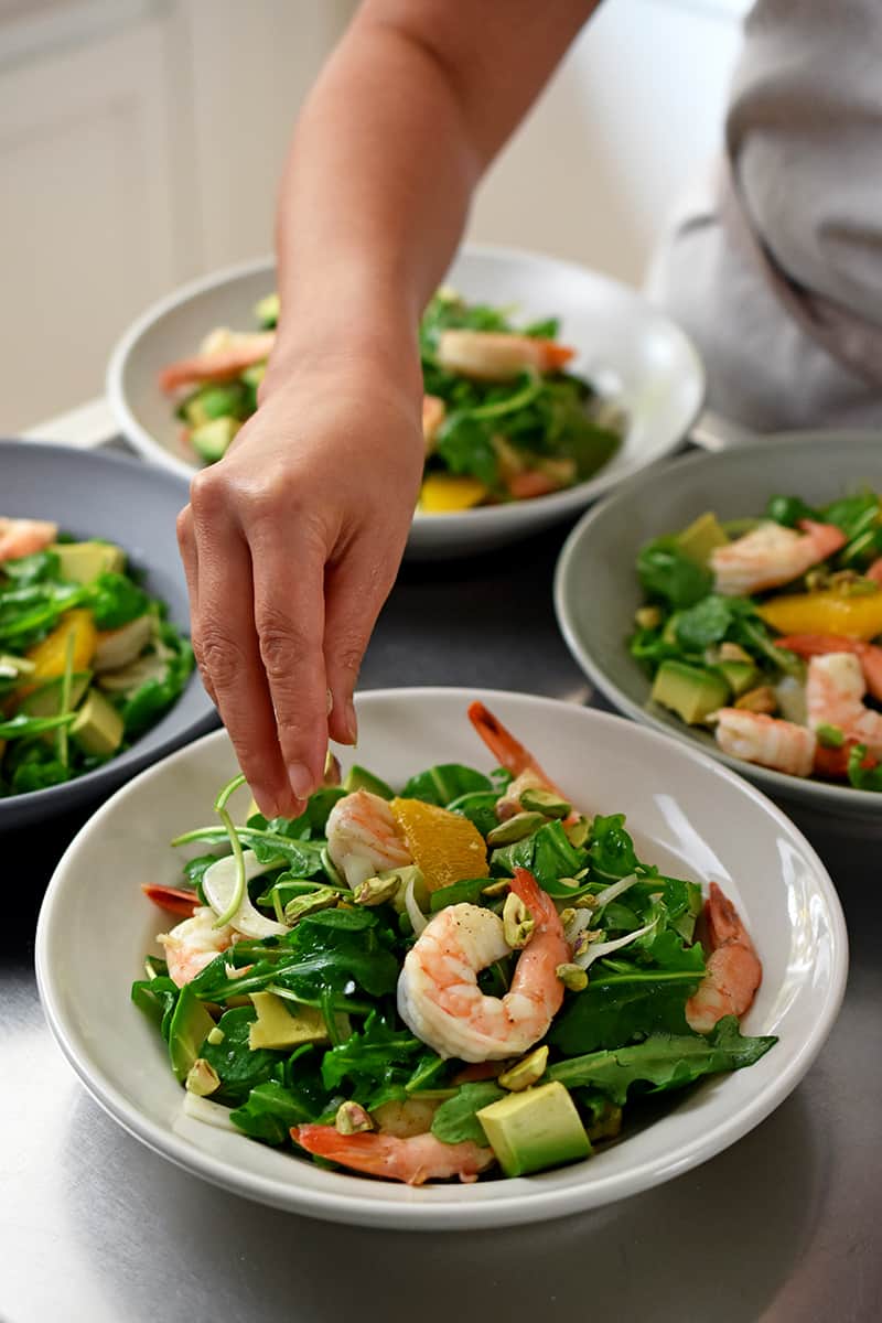 A hand is adding pistachios to a shrimp avocado salad.
