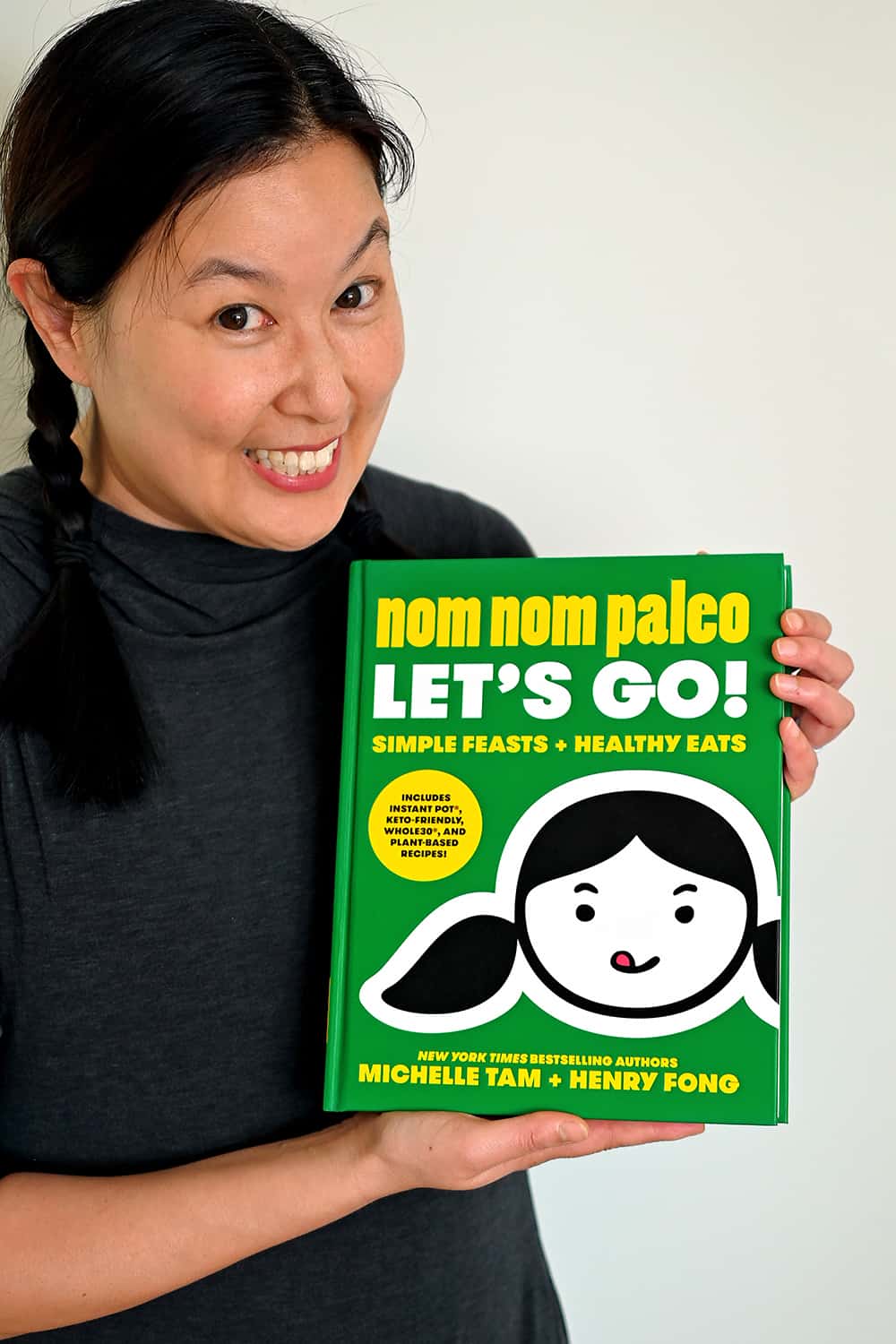 A smiling Asian woman is holding a green cookbook titled, 