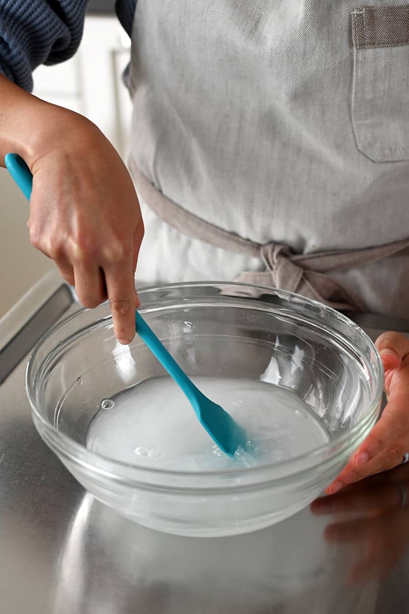 A blue silicone spatula is stirring a salt solution in a clear bowl.