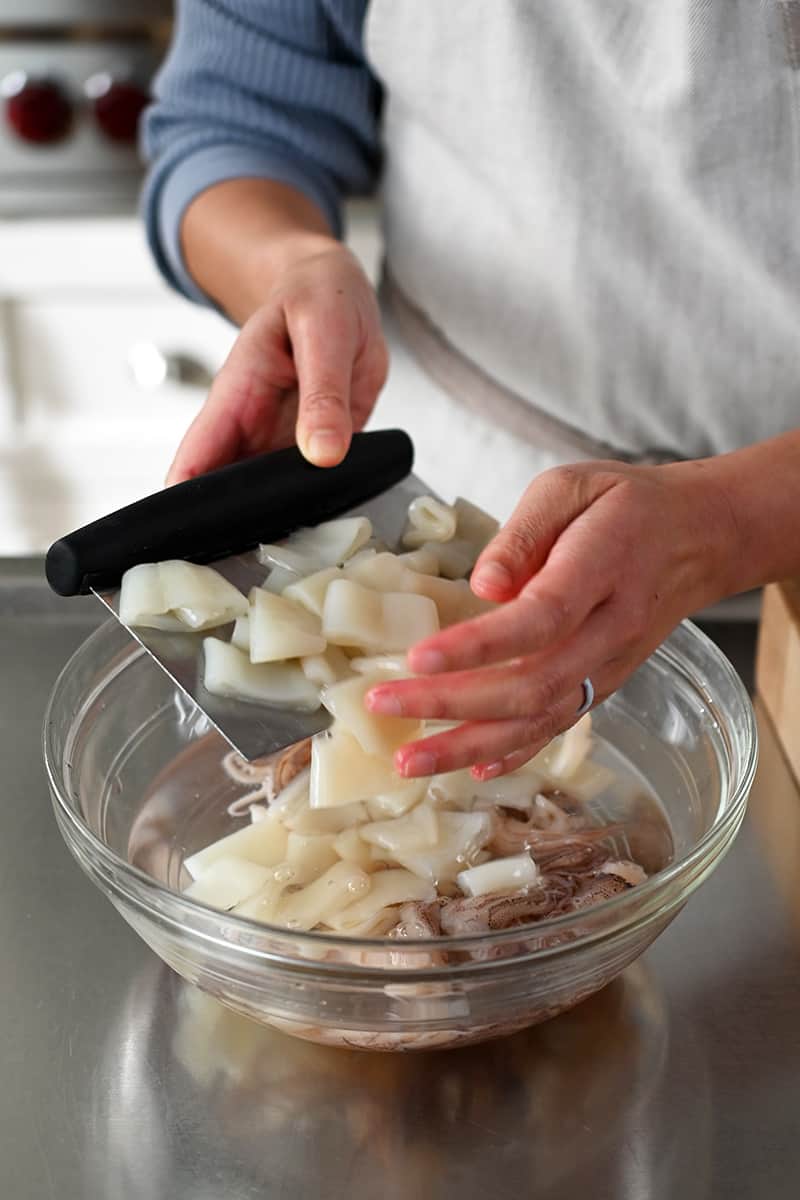 Two hands are adding sliced calamari to a clear bowl filled with a clear liquid.