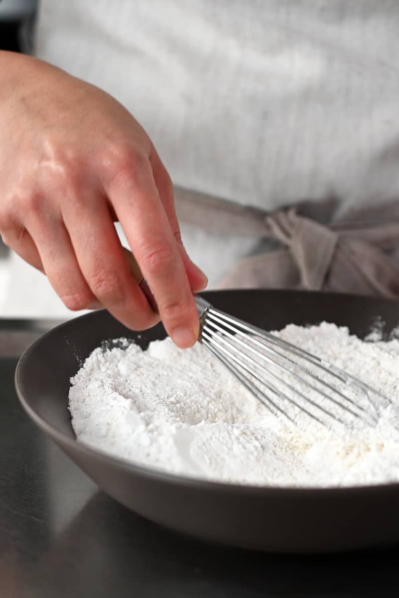 A hand is using a whisk to mix flour in a shallow brown bowl.