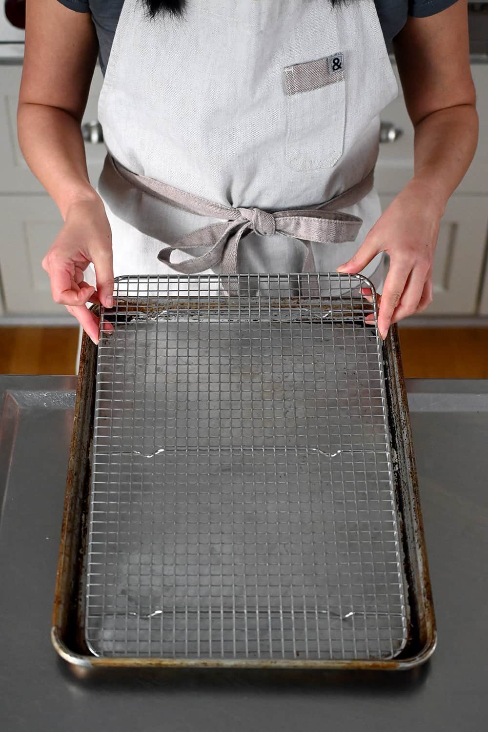 A person in an off-white apron is placing a wire rack inside a rimmed baking sheet