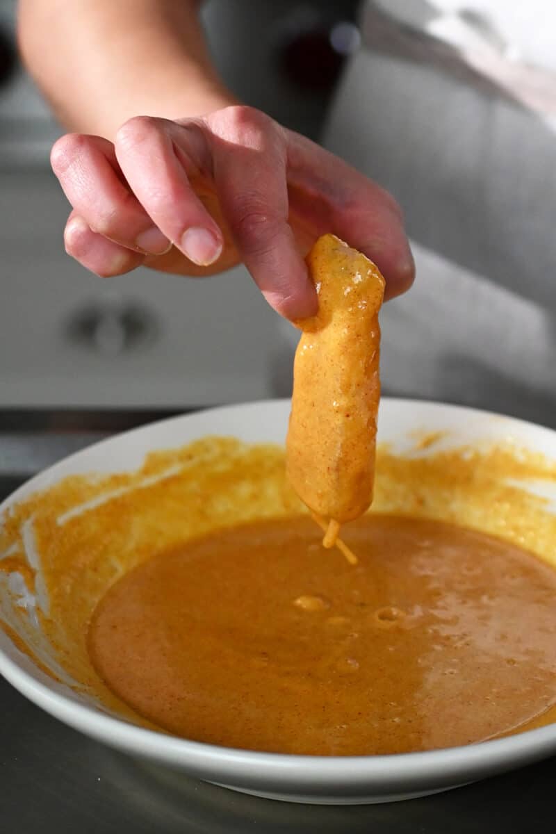 A hand is dipping a fish fillet into a bowl with seasoned egg and flour batter