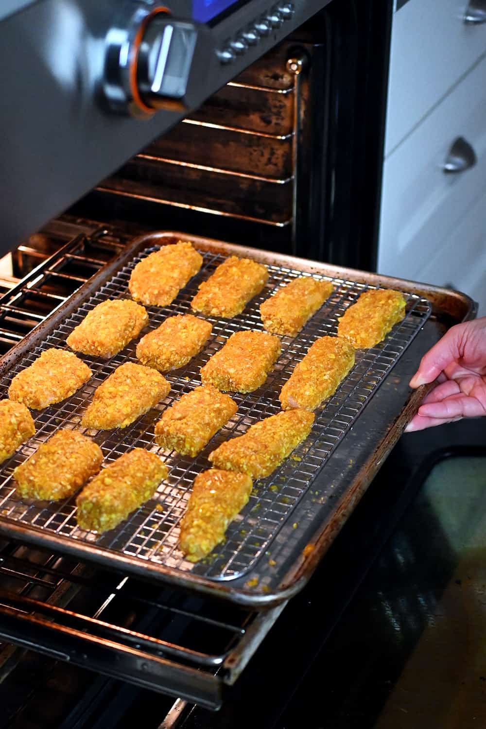 A hand is placing a tray of paleo fish sticks on a wire rack in a rimmed baking sheet into an oven,
