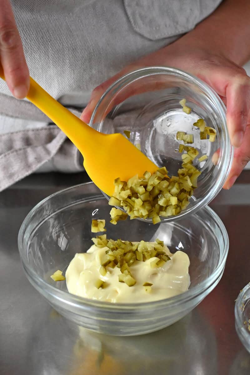 Adding diced pickles to a glass bowl filled with mayonnaise.