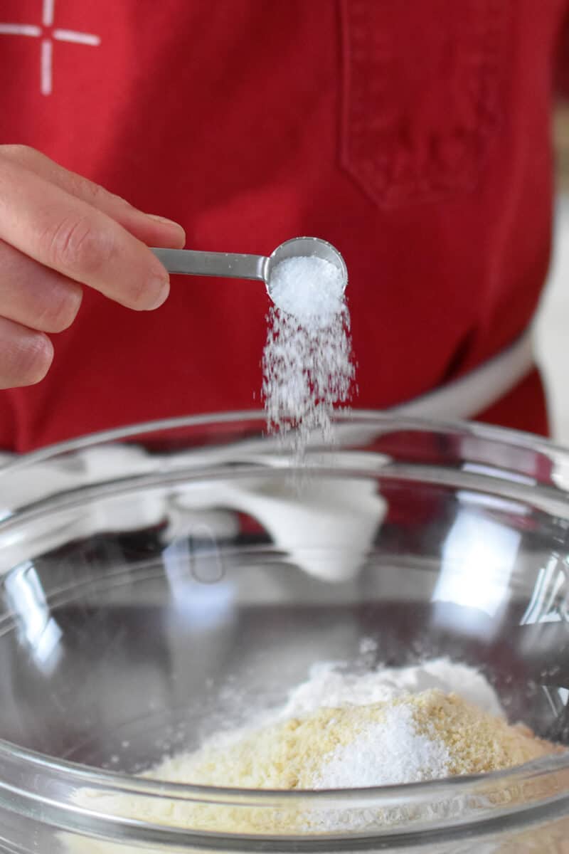 Adding a spoonful of salt to a glass bowl filled with the dry ingredients for paleo financiers