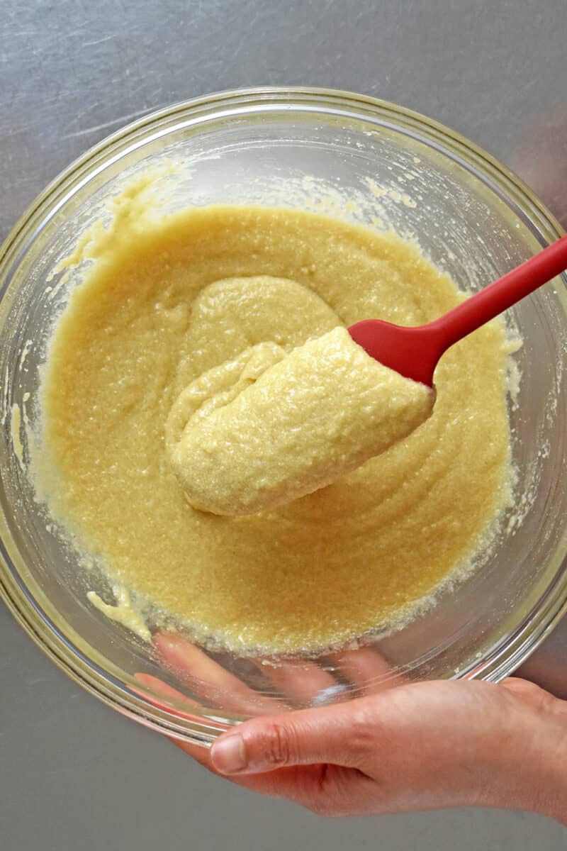 A red spatula is mixing the financiers batter in a glass mixing bowl.
