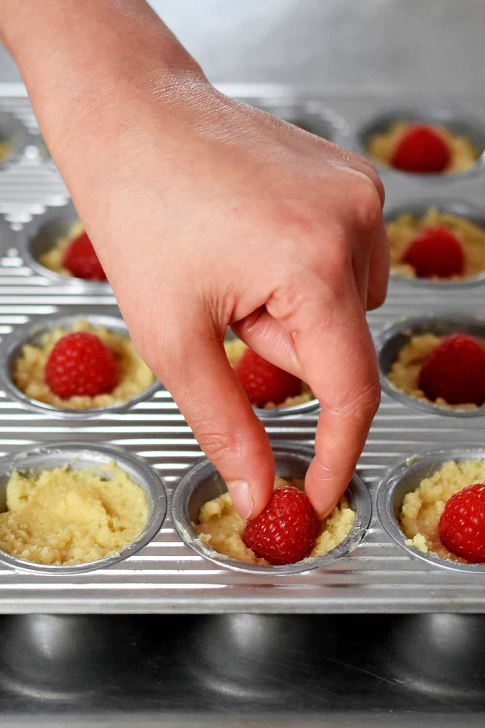 A hand is placing a raspberry into financier batter in a mini muffin pan
