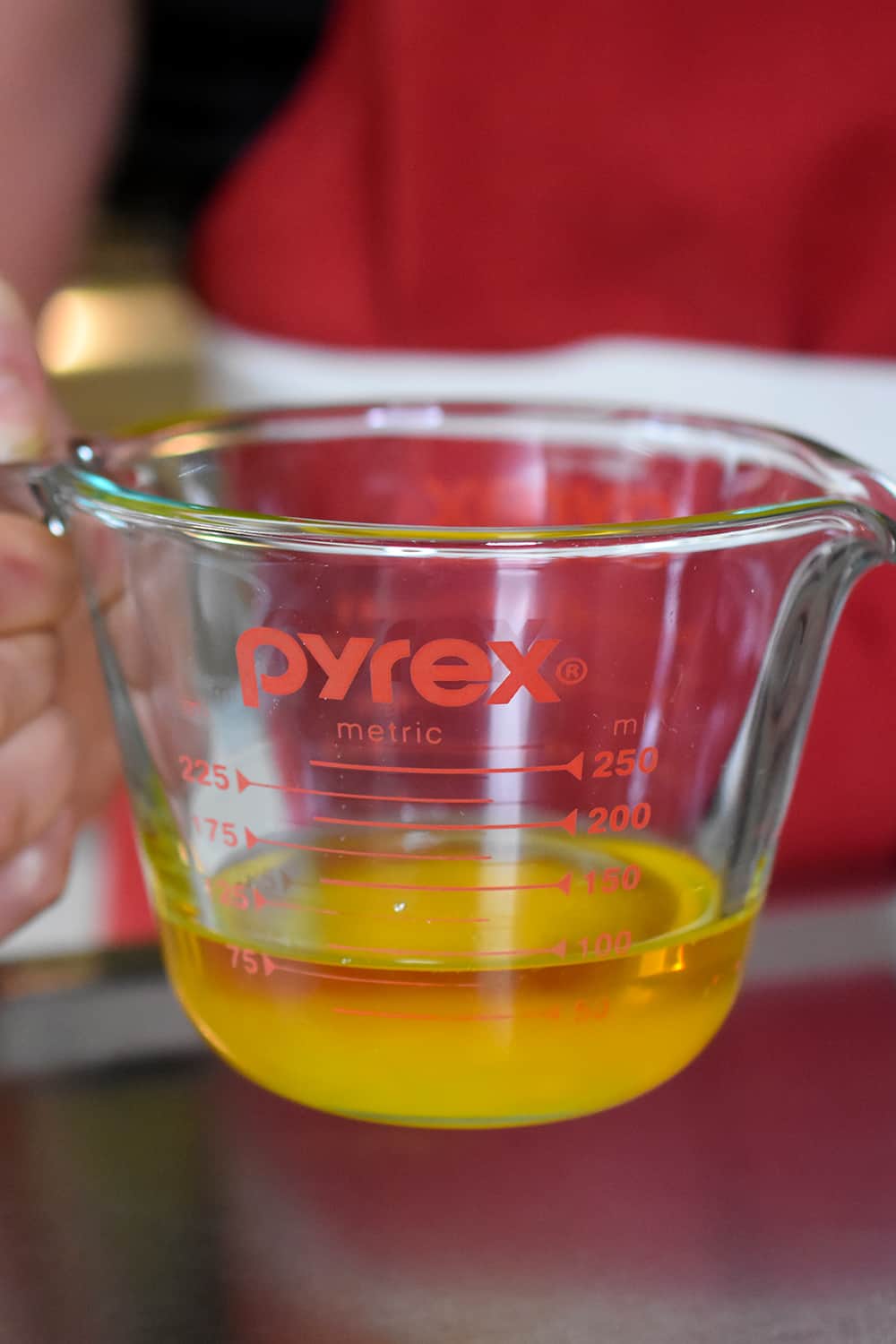 A hand is holding a liquid measuring cup with melted ghee inside.