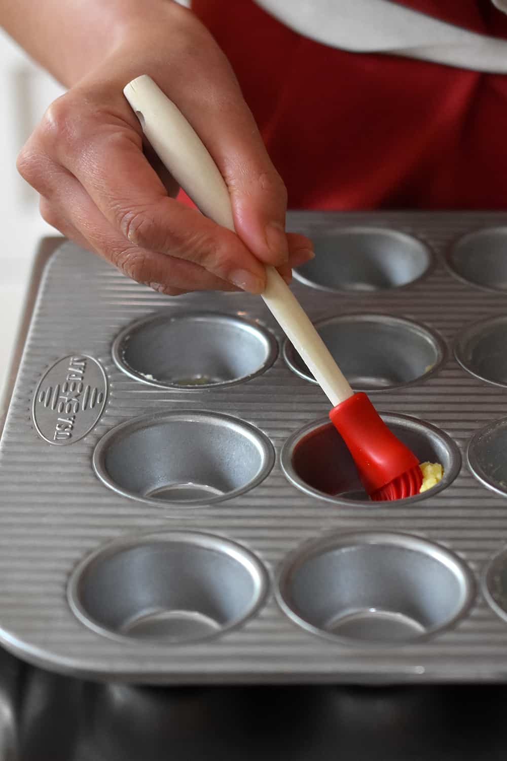 A hand is using a silicone brush to add ghee to grease a mini muffin tin.