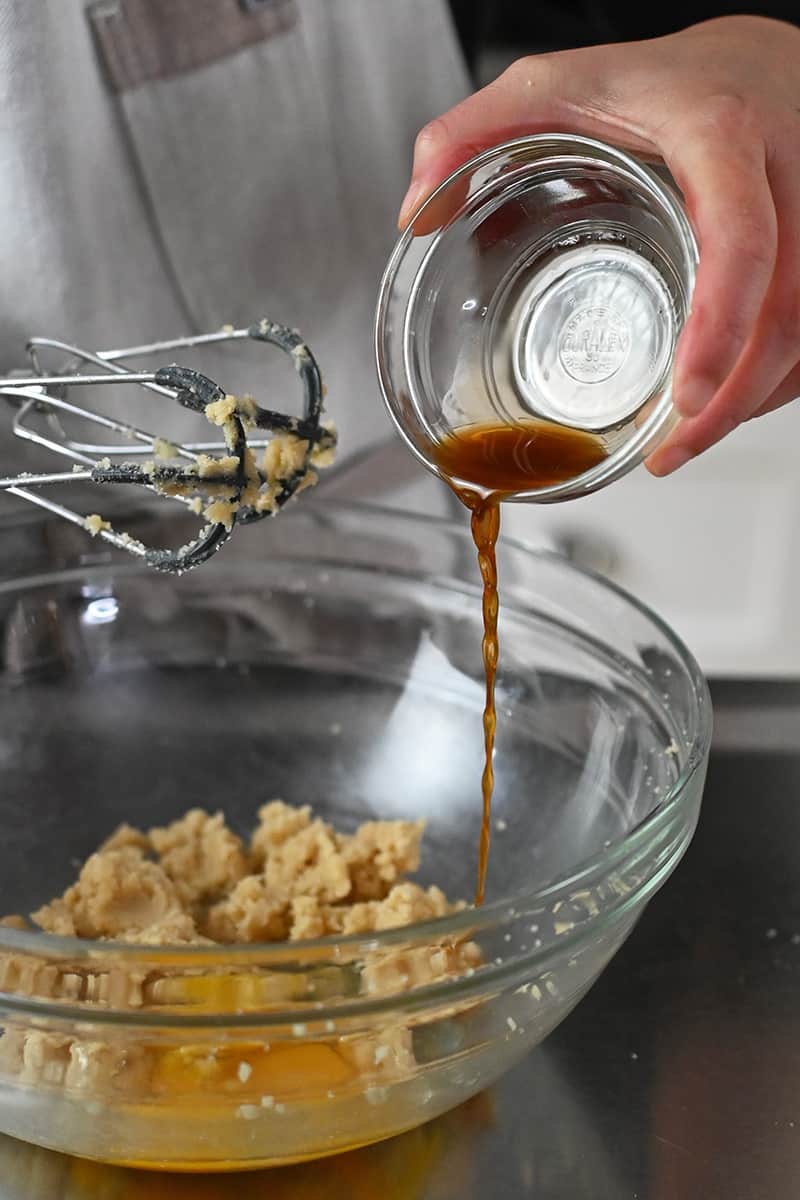 Pouring vanilla extract into a bowl with creamed sugar and ghee and a raw egg.