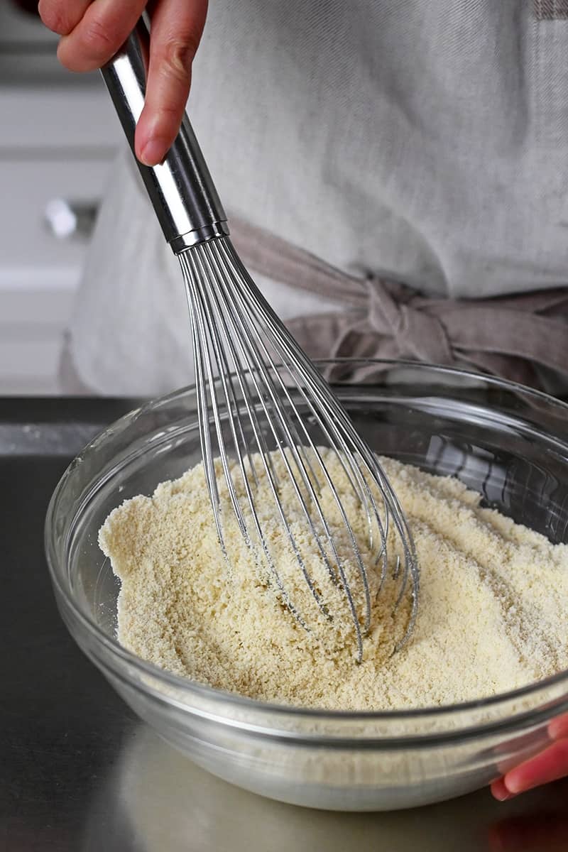 A whisk is combining the dry ingredients for paleo thumbprint cookies in a glass bowl.