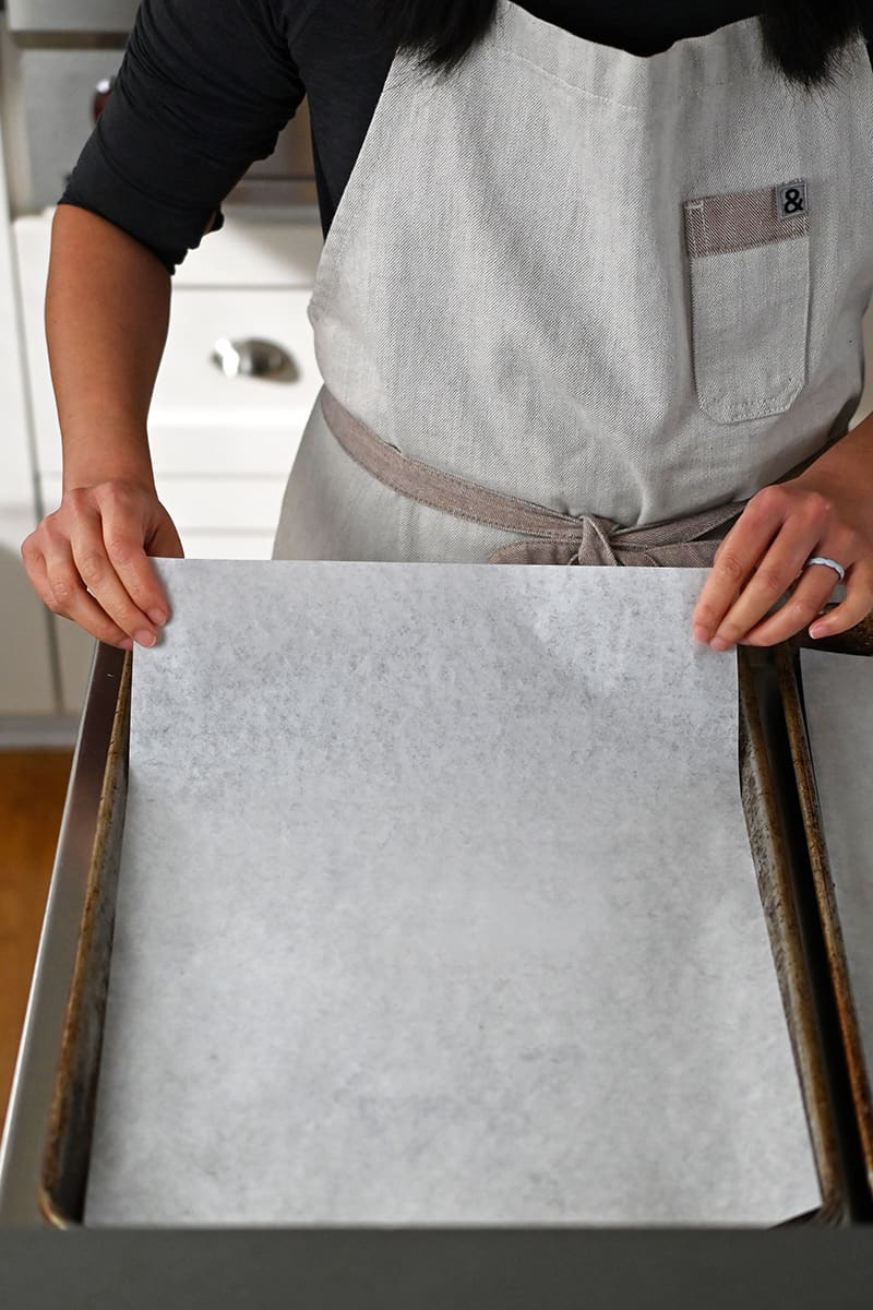 A person in an apron is putting a piece of parchment paper in a rimmed baking sheet