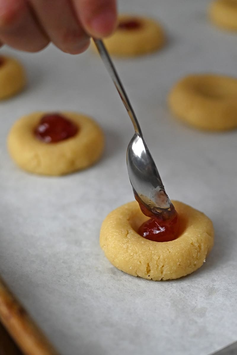 A small spoon is adding jam to the center of thumbprint cookie dough.