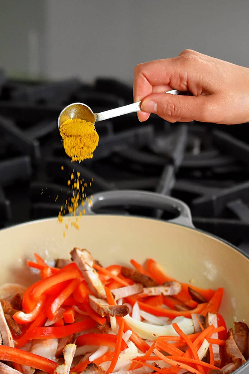 Someone adding a spoonful of curry powder to a skillet filled with matchstick vegetables, shrimp, and Char Siu