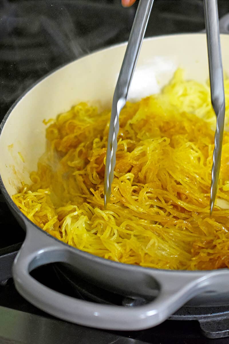 A pair of metal tongs is stirring spaghetti squash in an enameled cast iron skillet.