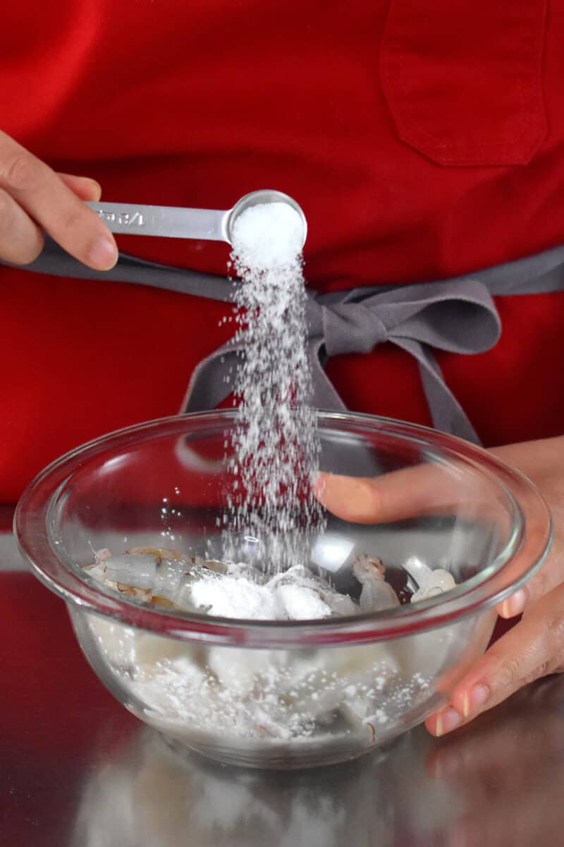 A person in a red apron is adding a spoonful of salt to a bowl of raw shrimp