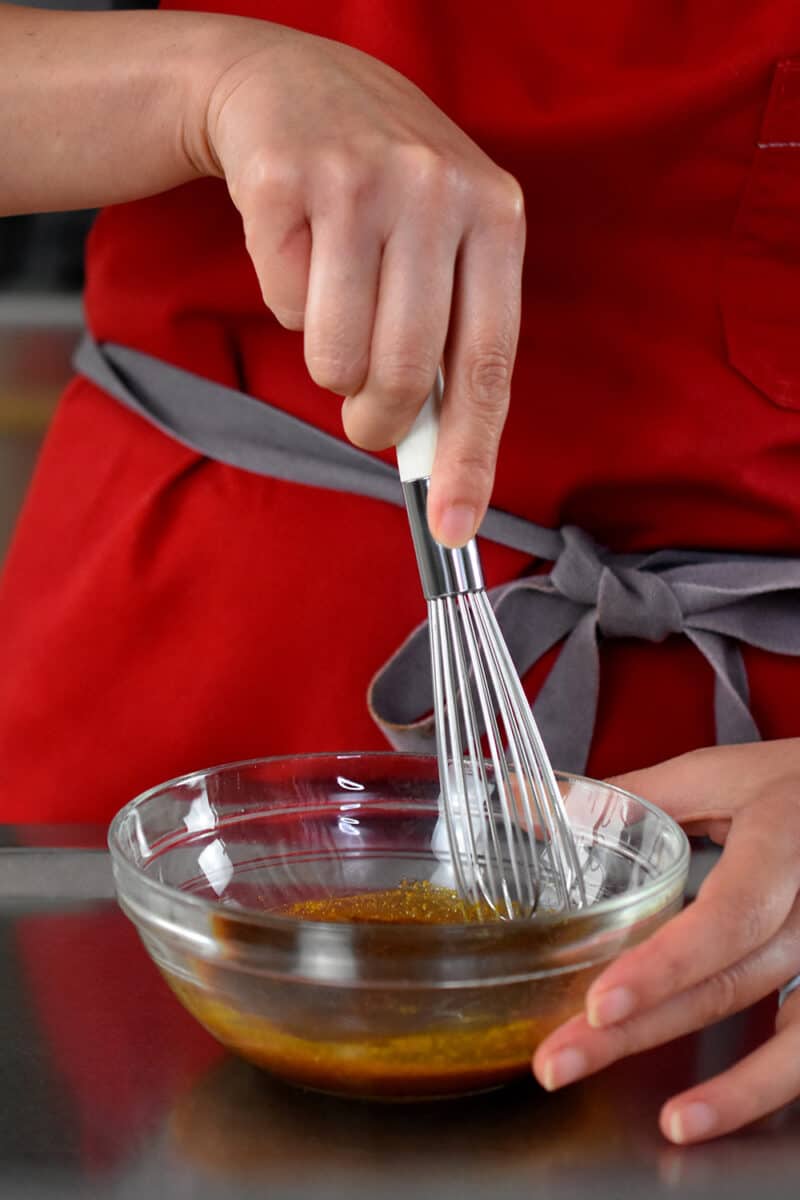 A person in a red apron is whisking the sauce for Singapore noodles in a small clear bowl