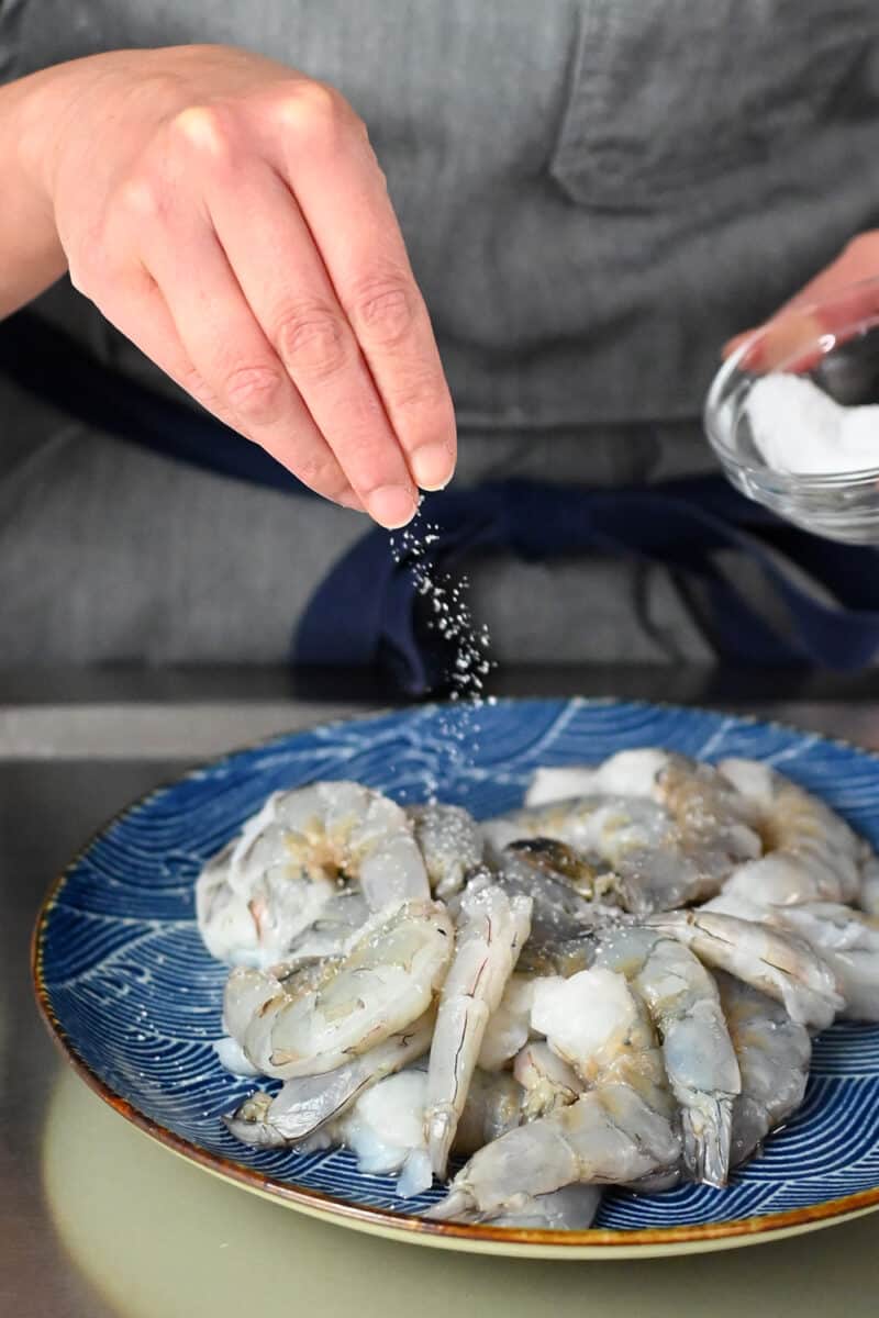 A hand is sprinkling salt on a plate of raw peeled jumbo shrimp
